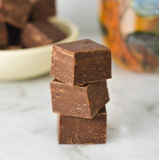 A stack of glossy Coffee Fudge, with more fudge squares and a coffee cup in the background.