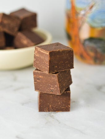A stack of glossy Coffee Fudge, with more fudge squares and a coffee cup in the background.