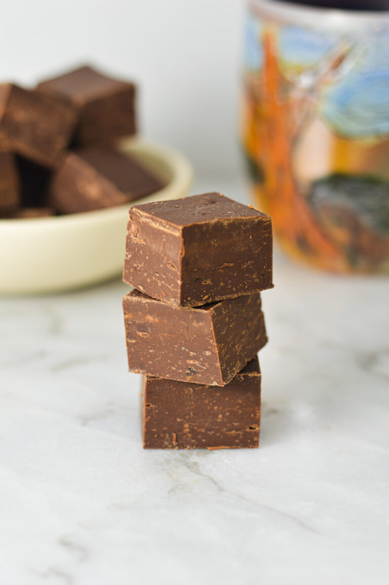A stack of glossy Coffee Fudge, with more fudge squares and a coffee cup in the background.
