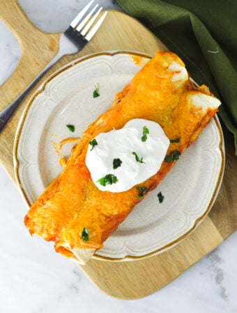 A plate with some Easy Leftover Turkey Enchiladas topped with melted Mexican blend cheese, sour cream and chopped cilantro.