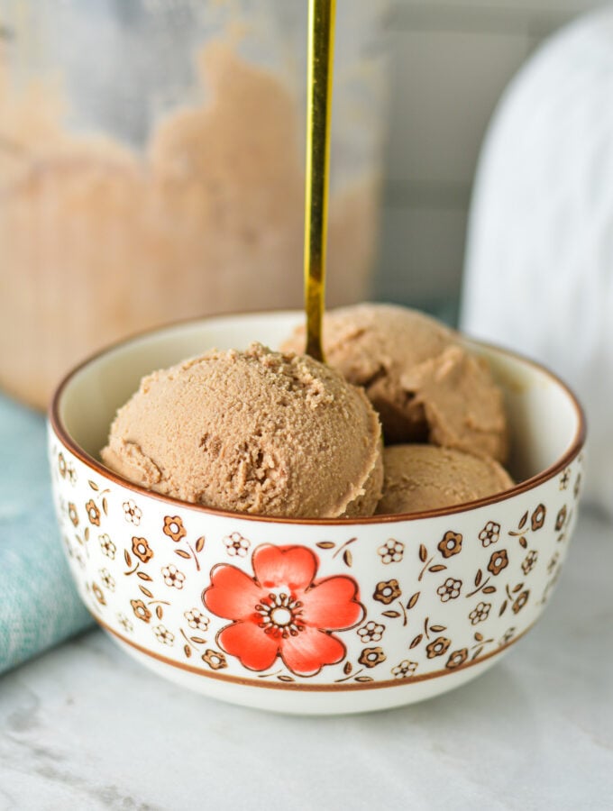 A gold spoon coming out of a bowl full of Ninja Creami Chocolate Ice Cream, with a Ninja Creami container in the background.