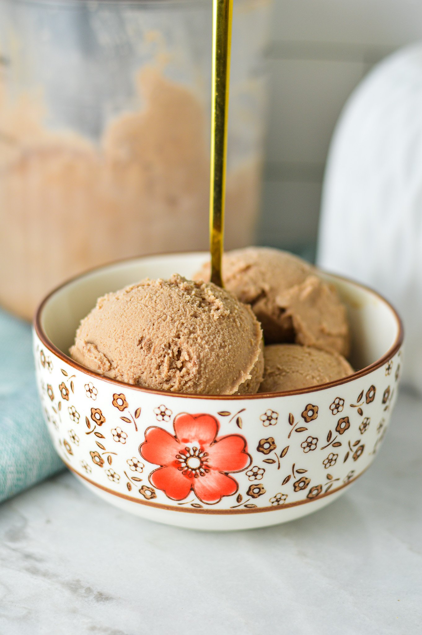 A gold spoon coming out of a bowl full of Ninja Creami Chocolate Ice Cream, with a Ninja Creami container in the background.