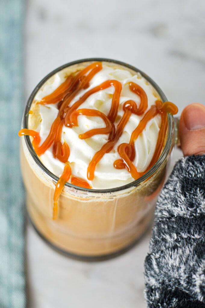 A hand holding a glass cup filled with Caramel Mocha and topped with whipped cream and a drizzle of caramel sauce, setting a cozy scene.