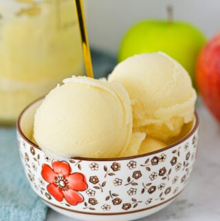 A bowl of Ninja Creami Apple Italian Ice with a gold spoon coming out of the icy treat, and apples in the background.