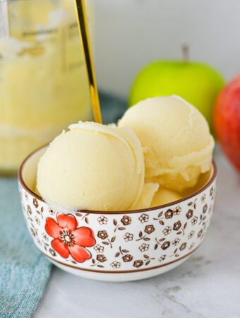 A bowl of Ninja Creami Apple Italian Ice with a gold spoon coming out of the icy treat, and apples in the background.