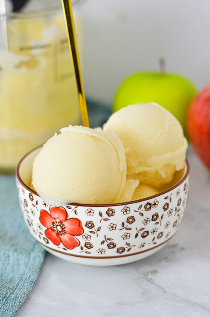 A bowl of Ninja Creami Apple Italian Ice with a gold spoon coming out of the icy treat, and apples in the background.