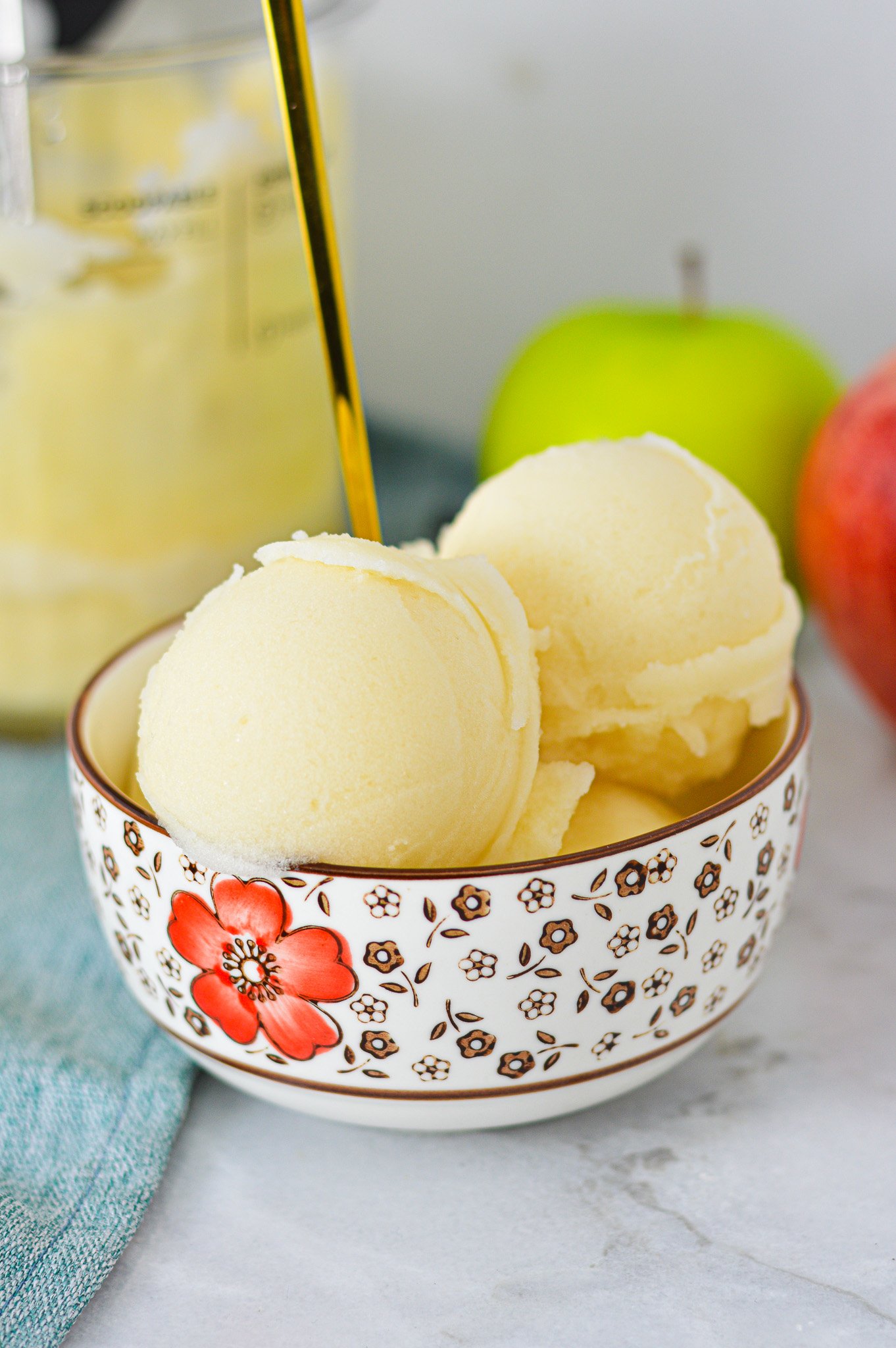 A bowl of Ninja Creami Apple Italian Ice with a gold spoon coming out of the icy treat, and apples in the background.