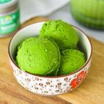 A small bowl filled with bright green Ninja Creami Matcha Ice Cream, with green tea powder in the background.