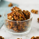 A small glass bowl filled with homemade Nutella Granola, with granola clumps on the granite surface in the background.