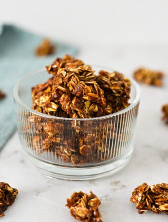 A small glass bowl filled with homemade Nutella Granola, with granola clumps on the granite surface in the background.
