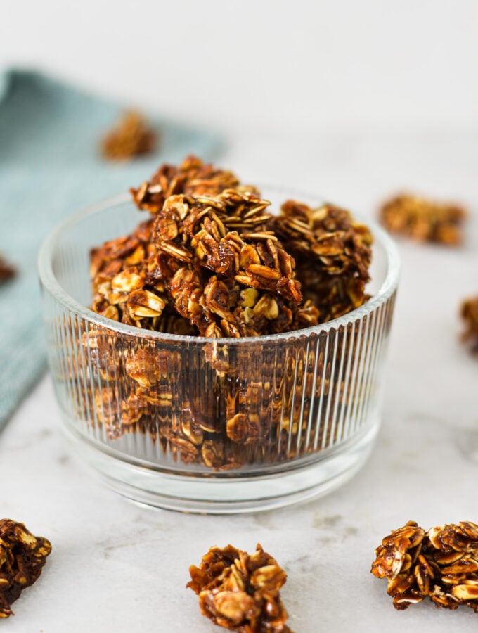 A small glass bowl filled with homemade Nutella Granola, with granola clumps on the granite surface in the background.