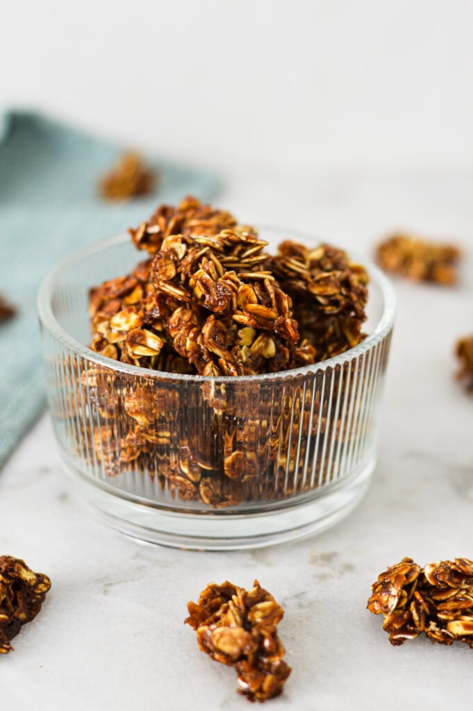 A small glass bowl filled with homemade Nutella Granola, with granola clumps on the granite surface in the background.