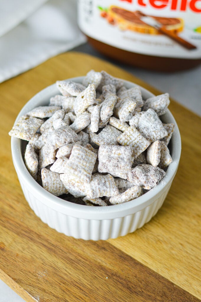 A ramekin filled with Nutella Puppy Chow, with each Chex cereal piece coated in Nutella chocolate and dusted with icing sugar.