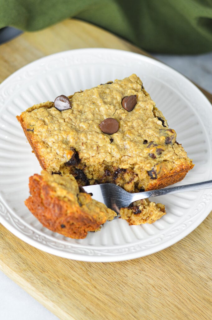 A square slice of Peanut Butter Chocolate Chip Baked Oatmeal on a small plate, with a fork taking out a piece of it to show the soft texture.
