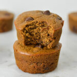 A stack of golden brown Protein Mocha Chocolate Chip Muffins with a bite taken out of the top muffin showing its soft texture.