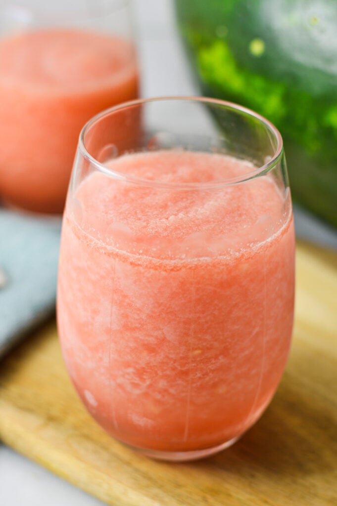 Two stemless wine glasses filled with Frozen Watermelon Daiquiri, with a slice of watermelon in the background.