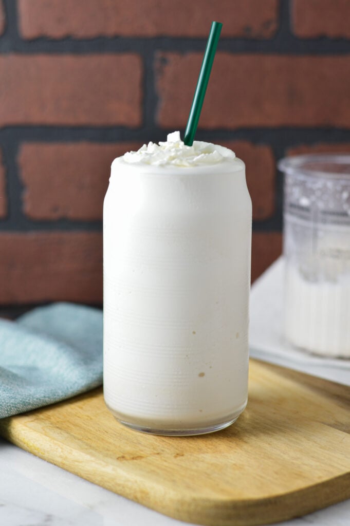 Creamy Ninja Creami Frozen White Hot Chocolate served in a clear glass, topped with whipped cream. A Ninja Creami pint is seen in the background.