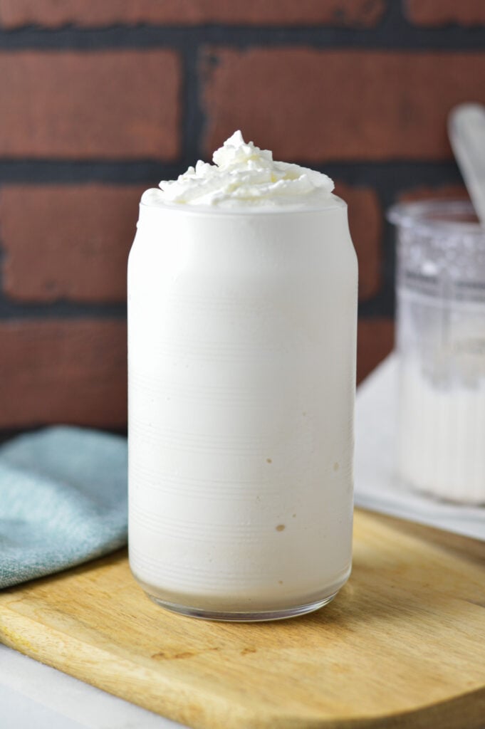 Refreshing Ninja Creami Frozen White Hot Chocolate served in a clear glass and topped with whipped cream. The pint from the Ninja Creami machine can be seen in the background.