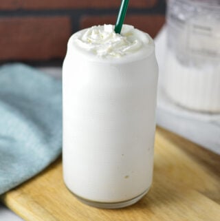 A tall glass of Ninja Creami Frozen White Hot Chocolate served on a wooden surface. The creamy dessert is topped with whipped cream, and a Ninja Creami container can be seen in the background.