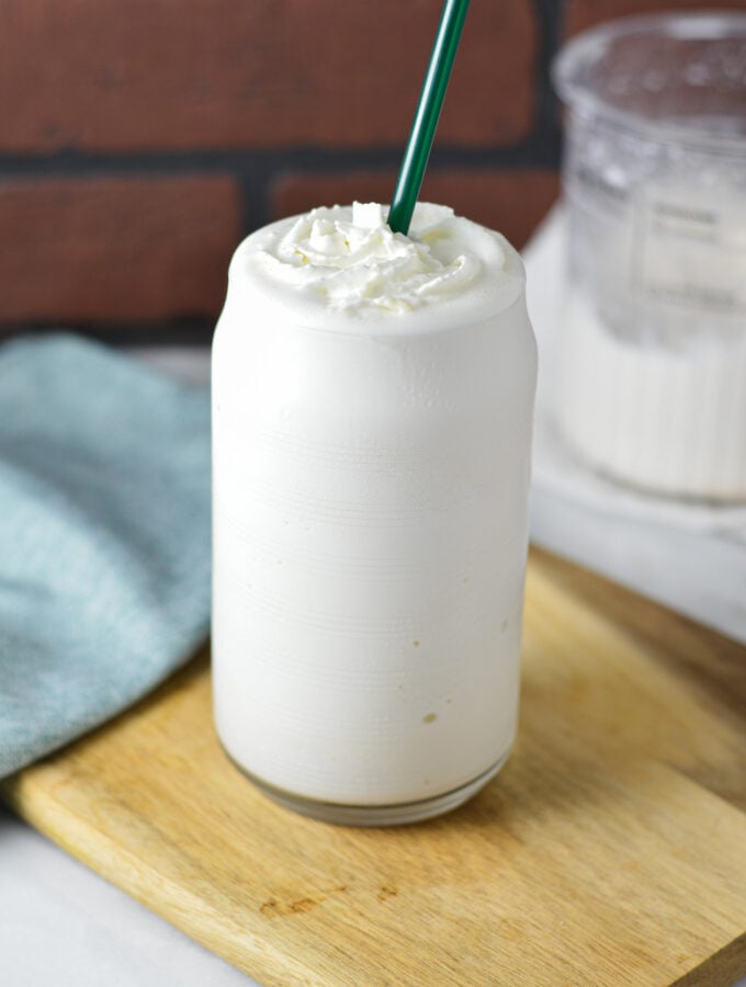 A tall glass of Ninja Creami Frozen White Hot Chocolate served on a wooden surface. The creamy dessert is topped with whipped cream, and a Ninja Creami container can be seen in the background.