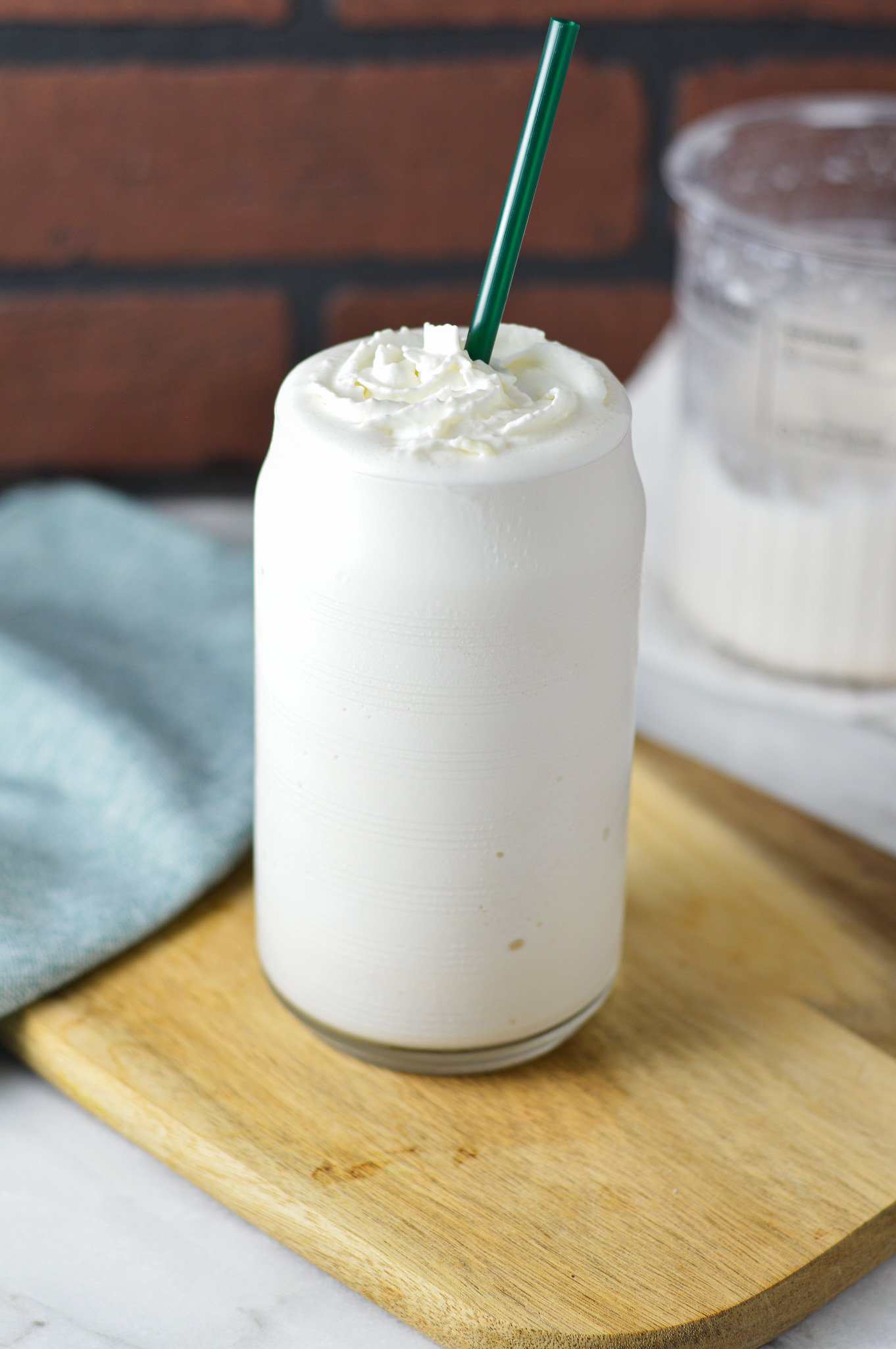 A tall glass of Ninja Creami Frozen White Hot Chocolate served on a wooden surface. The creamy dessert is topped with whipped cream, and a Ninja Creami container can be seen in the background.