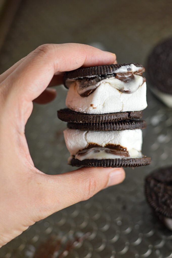 AA hand holding up two S'moreos, showing the gooey marshmallows and melted chocolate coming out from the two Oreo cookies.
