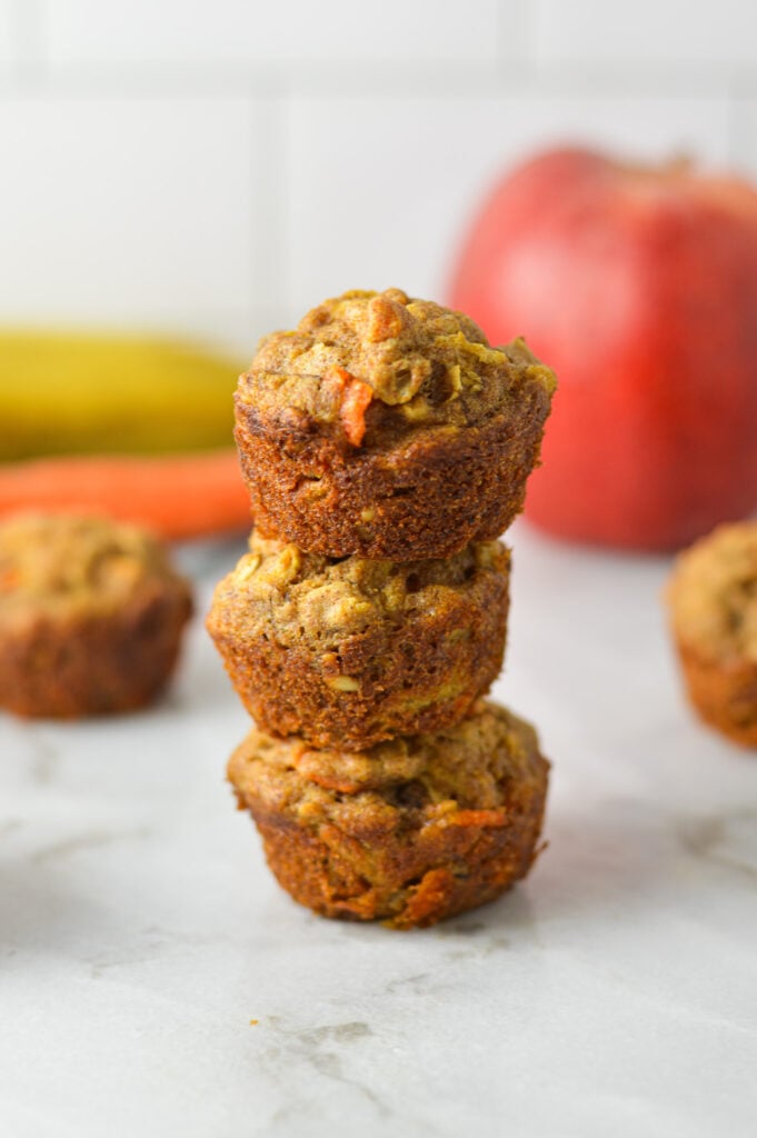 A stack of ABC Baby Mini Muffins, with an apple, an orange and a carrot in the background.