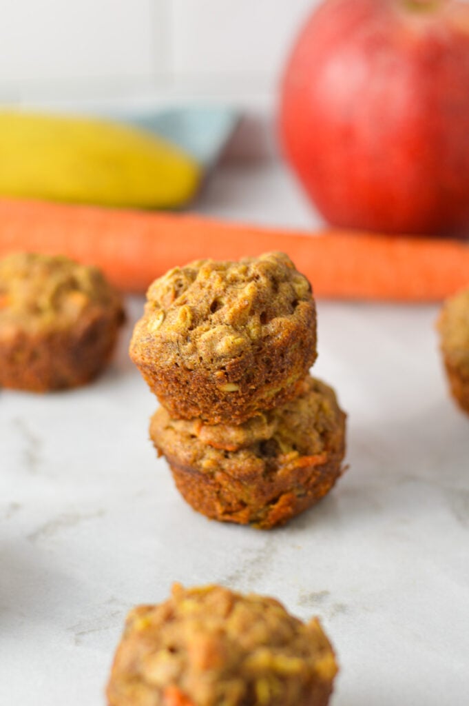 A stack of ABC Baby Mini Muffins on a granite surface, with some fruit and veggies in the background.