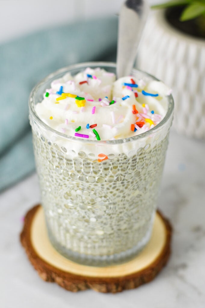 A spoon in a cup full of birthday cake flavored chia pudding, topped with whipped cream and multicolored sprinkles.