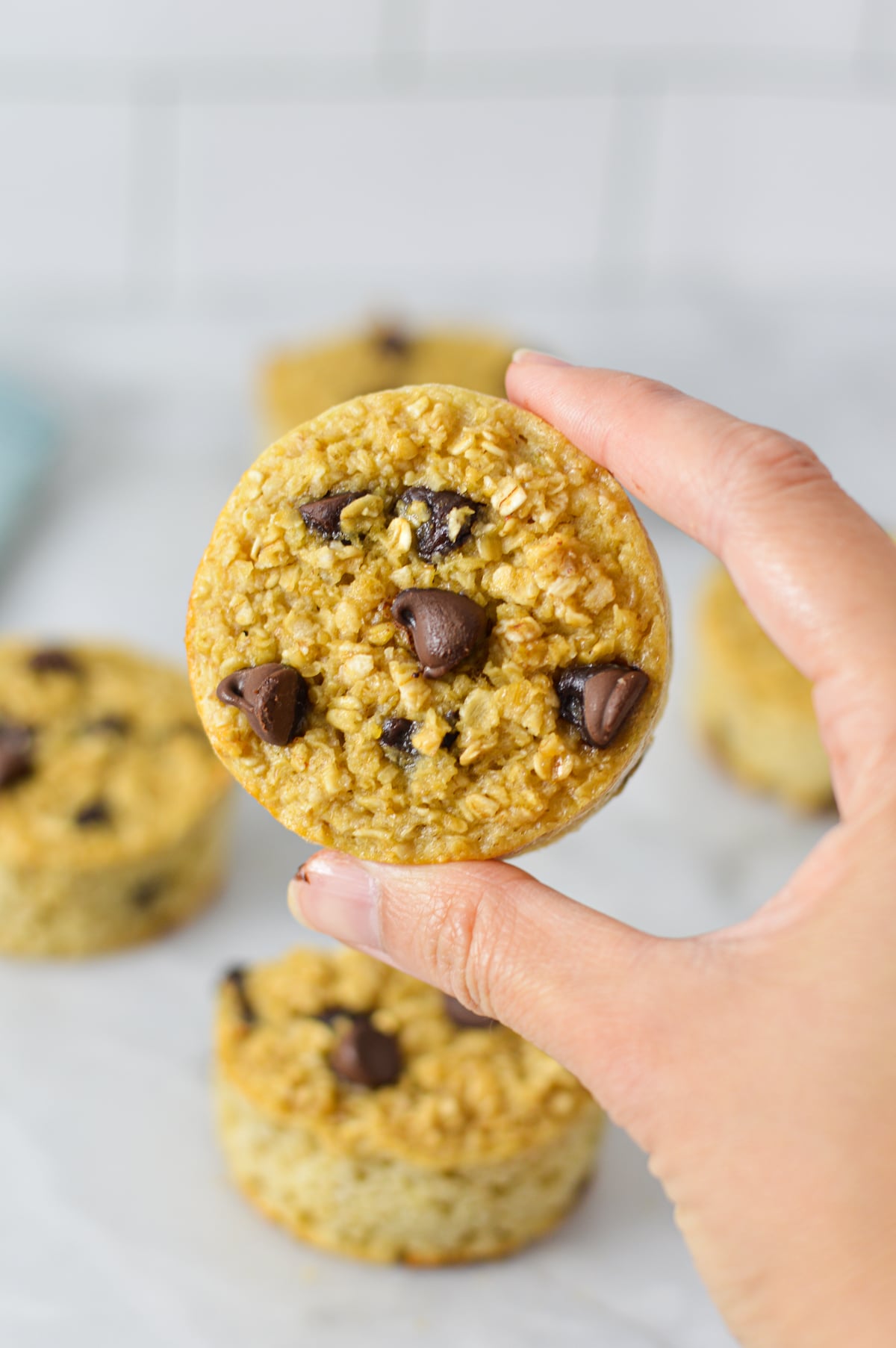 A hand holding homemade Chocolate Chip Baked Oatmeal Cups, with more freshly baked oatmeal cups in the background.