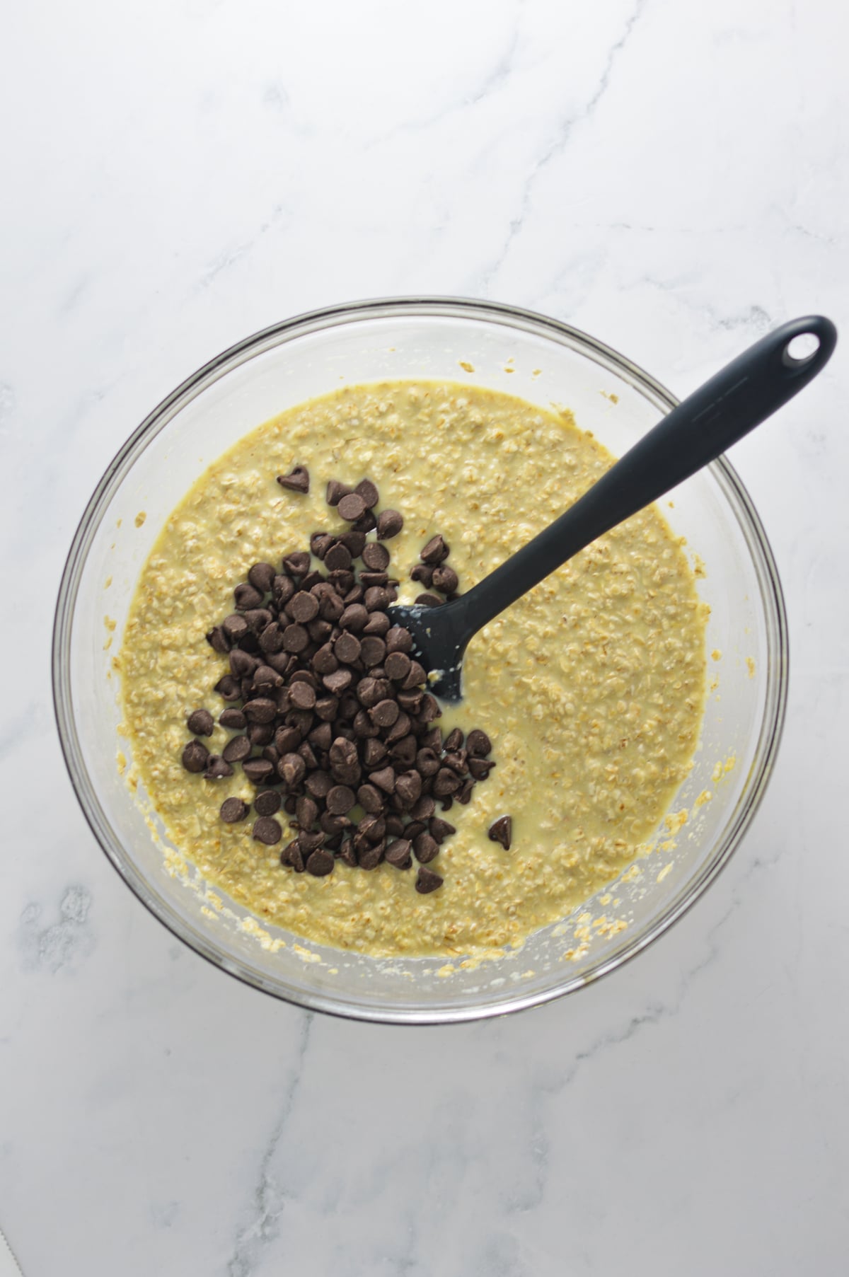 A glass bowl filled with Chocolate Chip Baked Oatmeal Cup batter, with semi-sweet chocolate chips on top and a rubber spatula ready to mix the batter.