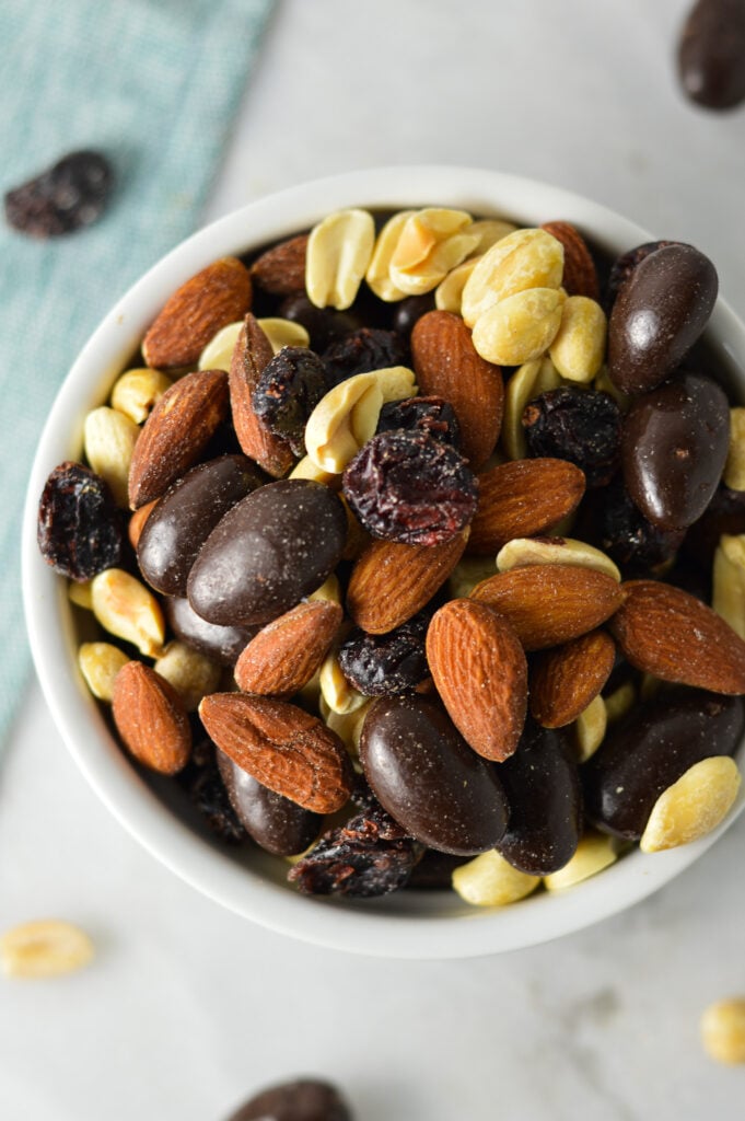 A small bowl full of Cranberry Chocolate Almond Trail Mix, with more of the homemade trail mix scattered on the granite surface in the background.