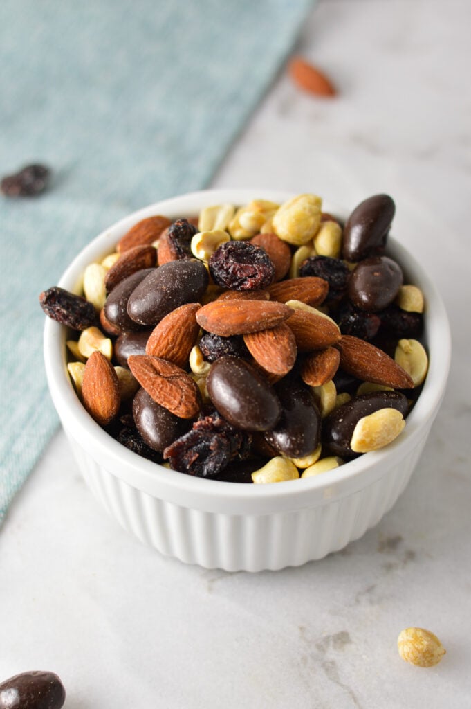 A white ramekin filled with Cranberry Chocolate Almond Trail Mix, with some more craisins and salted peanuts in the background.