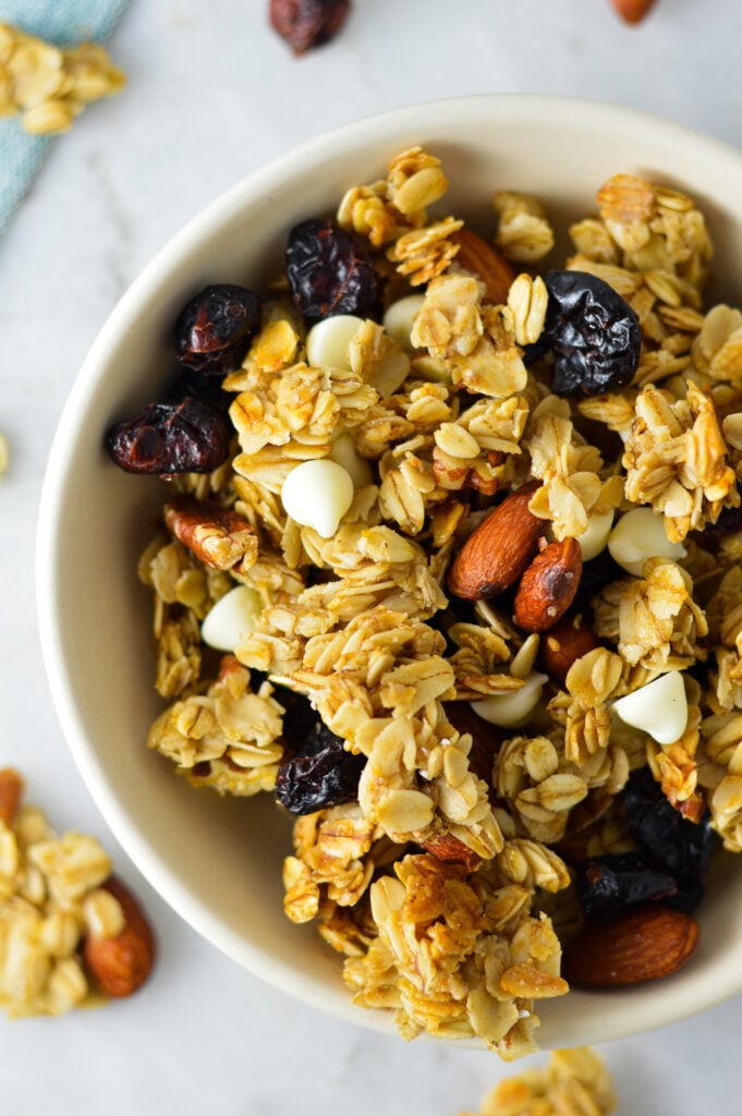A white bowl full of homemade Cranberry White Chocolate Granola, with chunks of granola dispersed on the granite surface.