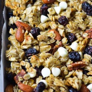 A parchment paper lined baking sheet with Cranberry White Chocolate Granola, with the white chocolate, whole almonds, pecans and cranberries evenly distributed in the homemade granola.