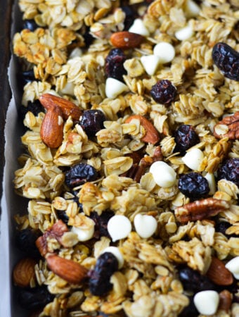 A parchment paper lined baking sheet with Cranberry White Chocolate Granola, with the white chocolate, whole almonds, pecans and cranberries evenly distributed in the homemade granola.