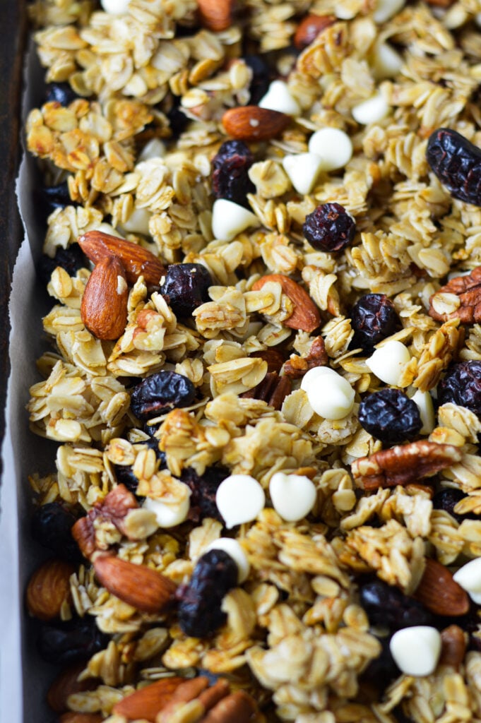 A parchment paper lined baking sheet with Cranberry White Chocolate Granola, with the white chocolate, whole almonds, pecans and cranberries evenly distributed in the homemade granola.