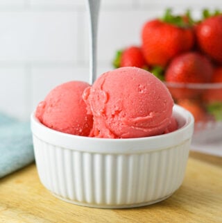 Large scoops of Ninja Creami Strawberry Sorbet in a small ramekin with a spoon sticking out of the homemade sorbet and fresh strawberries in the background.