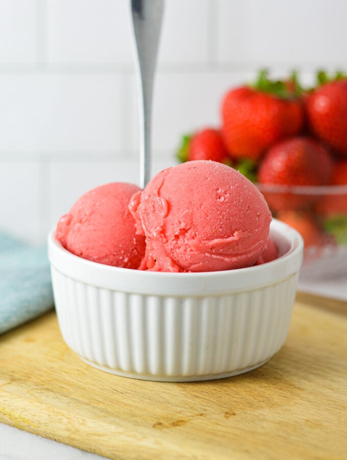 Large scoops of Ninja Creami Strawberry Sorbet in a small ramekin with a spoon sticking out of the homemade sorbet and fresh strawberries in the background.
