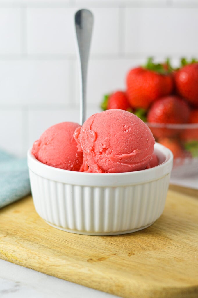 Large scoops of Ninja Creami Strawberry Sorbet in a small ramekin with a spoon sticking out of the homemade sorbet and fresh strawberries in the background.