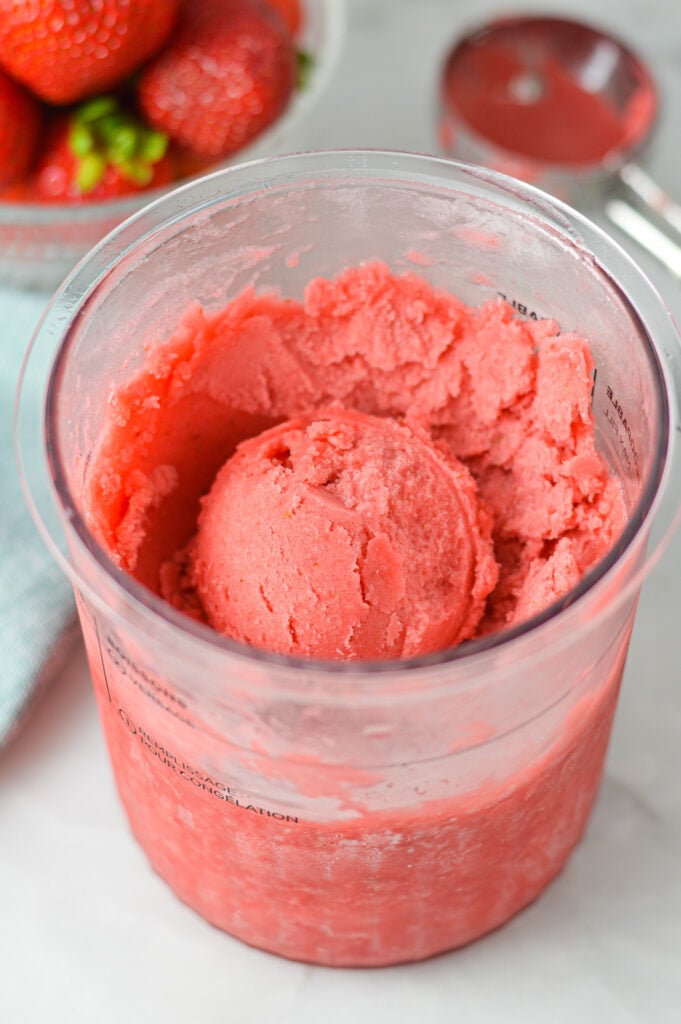 A Ninja Creami Pint container filled with Ninja Creami Strawberry Sorbet, with a large ice cream scoop and ripe strawberries in the background.
