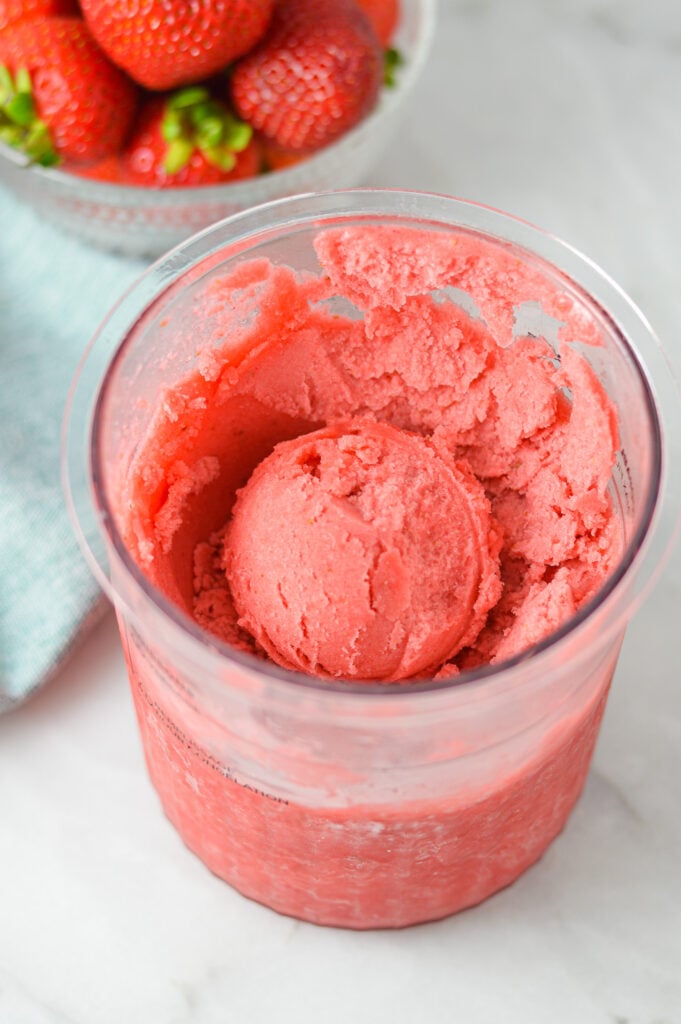 A Ninja Creami container filled with homemade Ninja Creami Strawberry Sorbet with a bowl full of fresh strawberries in the background.