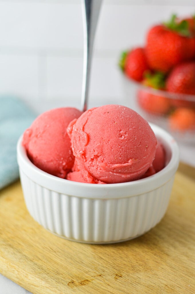 A white ramekin filled with scoops of Ninja Creami Strawberry Sorbet with a small spoon sticking out of the sorbet and fresh strawberries in the background.