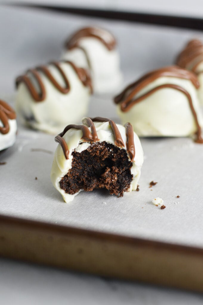 Some Nutella White Chocolate Cake Balls on a parchment paper lined baking tray, with a bite taken out of the front cake ball showing the fudgy texture inside.