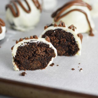 Some Nutella White Chocolate Cake Balls on a cookie sheet covered in wax paper. One cake ball is cut in half, showing the chocolate cake inside of the cake ball.