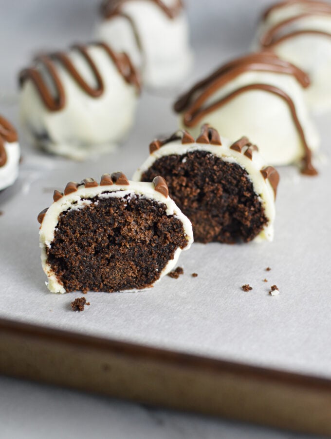 Some Nutella White Chocolate Cake Balls on a cookie sheet covered in wax paper. One cake ball is cut in half, showing the chocolate cake inside of the cake ball.