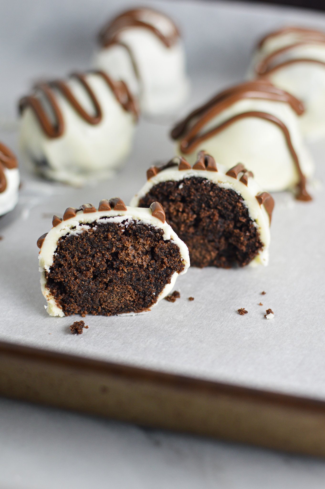 Some Nutella White Chocolate Cake Balls on a cookie sheet covered in wax paper. One cake ball is cut in half, showing the chocolate cake inside of the cake ball.