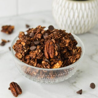 A bowl of homemade Chocolate Granola, with some pecans and chocolate chips on the granite surface.