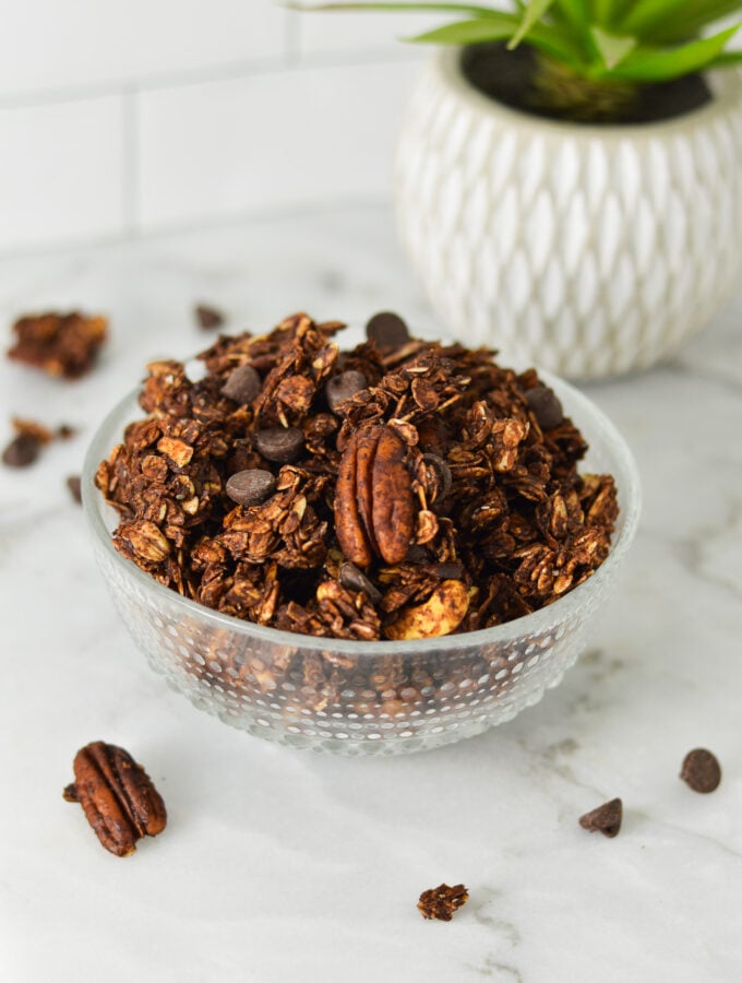 A bowl of homemade Chocolate Granola, with some pecans and chocolate chips on the granite surface.