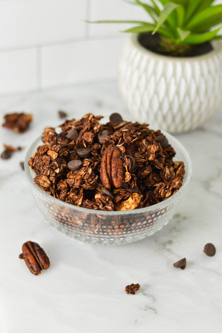 A bowl of homemade Chocolate Granola, with some pecans and chocolate chips on the granite surface.